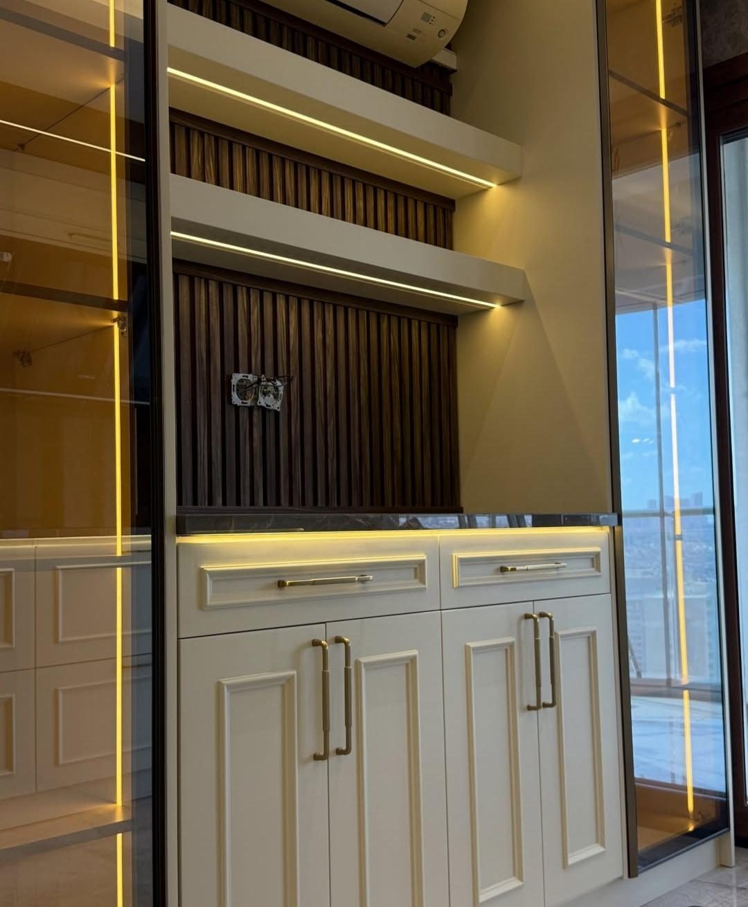 Kanallı ceviz panelli salon ünitesi, LED aydınlatmalı raflar ve pirinç kulplu krem dolaplar