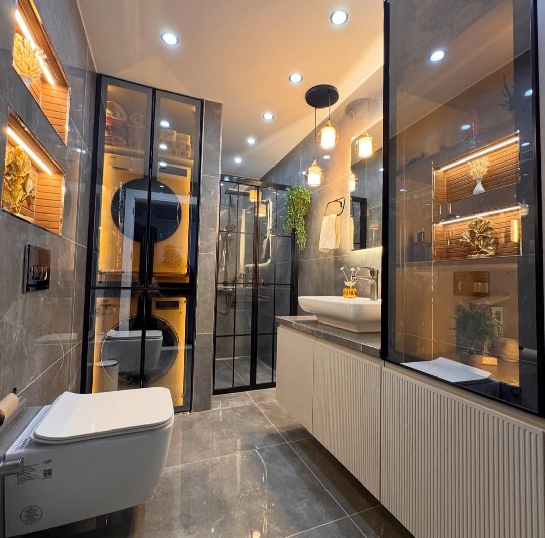 Geniş banyoda siyah çerçeveli camlı vitrin kolonları, kanallı lavabo dolabı ve sarkıt aydınlatma